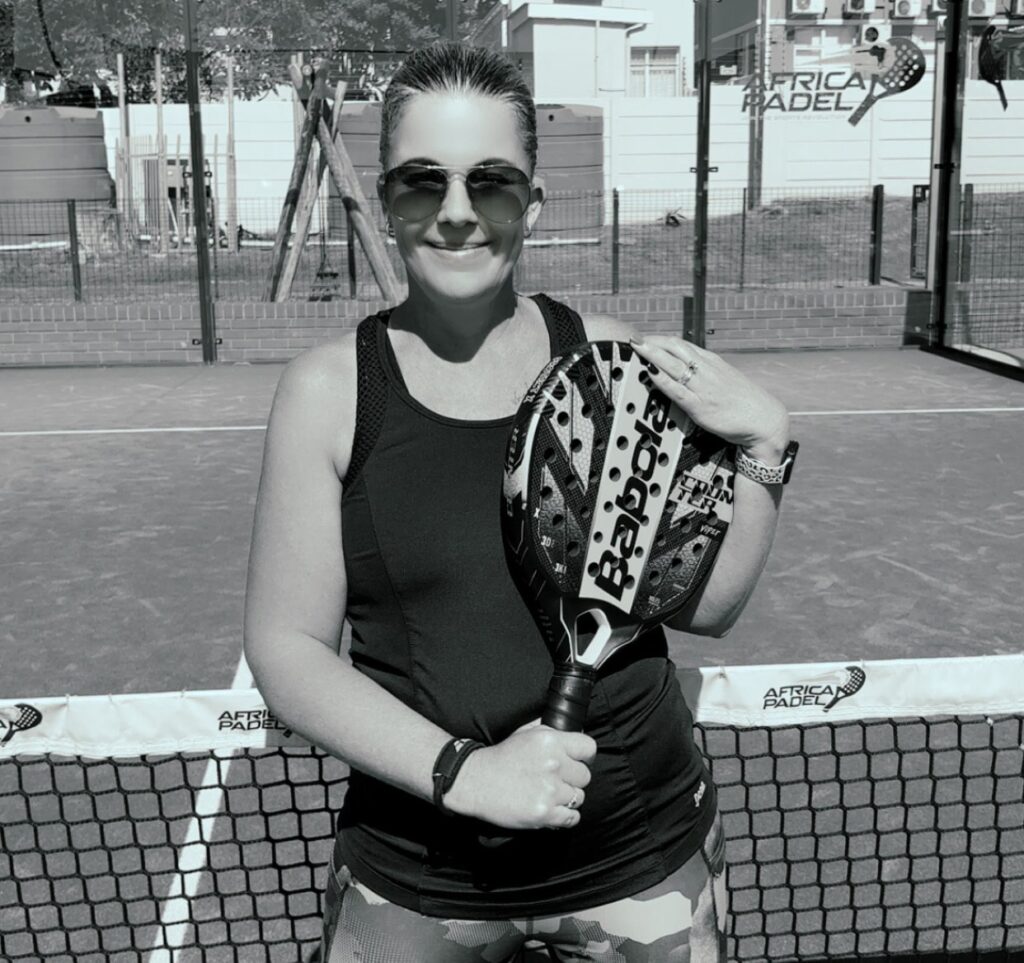 Padel player wearing sunglasses and camouflage leggings holding a Babolat racket on an outdoor padel court, with net branding and urban background.