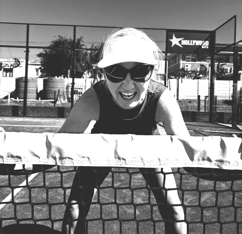 Black and white image of padel player in visor and sunglasses leaning on net, with urban backdrop, capturing intensity and style.