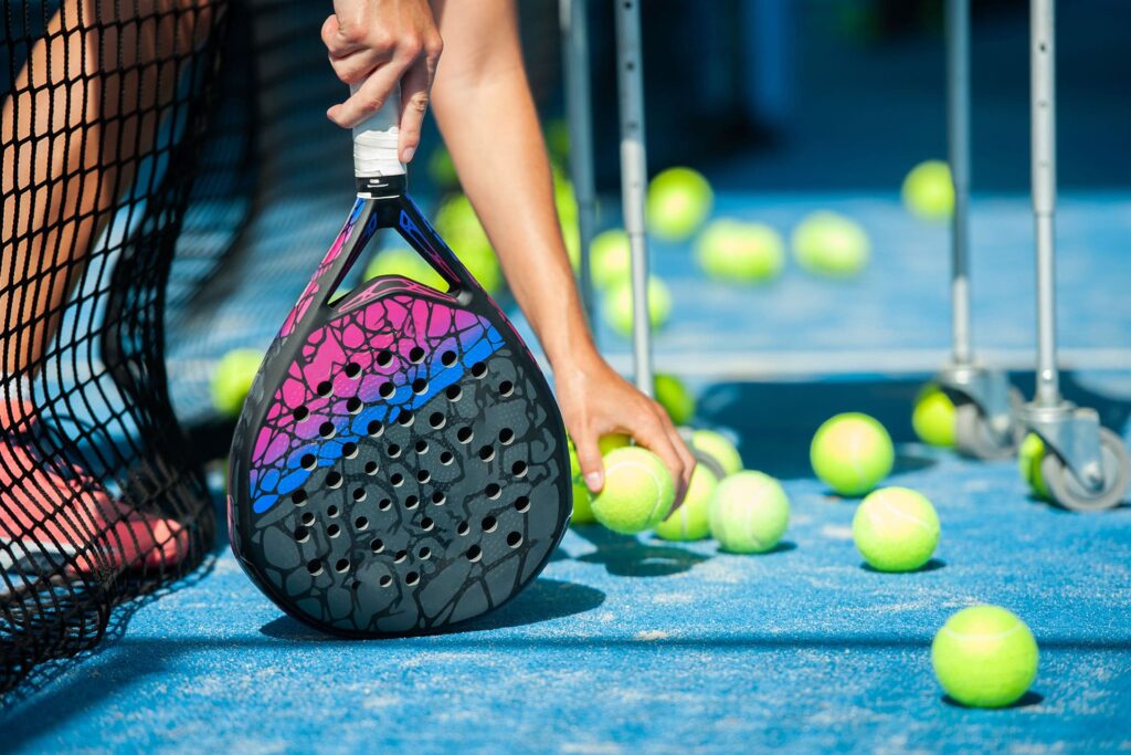Close-up of padel player picking up tennis balls on a blue padel court, holding a colorful perforated padel racket. Training gear and ball cart visible, emphasizing padel practice and equipment setup.